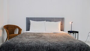 A neatly made bed with a grey blanket, a wicker chair, and a bedside table with a lamp. - Arecibo Hotel 2104 (Arecibo)