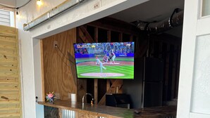 An outdoor bar area with a television displaying a baseball game, wooden stools, and a metal countertop. - Canal House, New Bar/TV Area w/ view of water, Fenced yard & 5 Min Walk To Beach (Ocean Isle Beach)