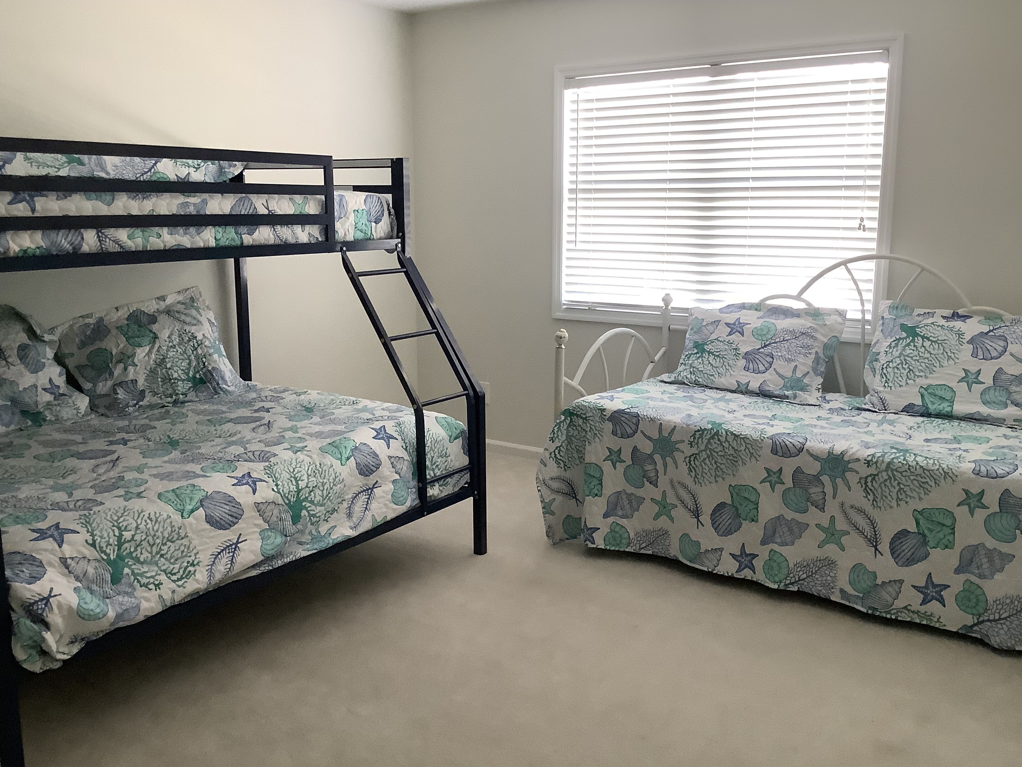 A bunk bed with a sea-themed bedspread and matching pillows, a window with blinds, and a white metal headboard.