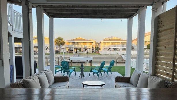 A covered outdoor seating area with a table and chairs, overlooking a pool and buildings. - Canal House, New Bar/TV Area w/ view of water, Fenced yard & 5 Min Walk To Beach (Ocean Isle Beach)