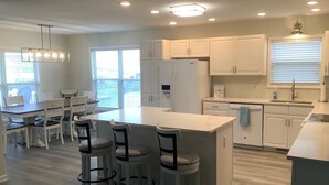 A modern kitchen with white cabinets, a central island with bar stools, and a dining area with a table and chairs. - Canal House, New Bar/TV Area w/ view of water, Fenced yard & 5 Min Walk To Beach (Ocean Isle Beach)