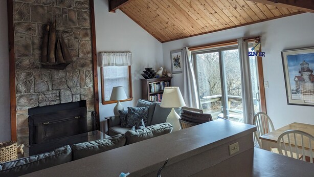 A cozy living room with a stone fireplace, wooden ceiling beams, a sofa, and a dining area with a table and chairs. - Private cottage in Madaket just a 10 min walk or short drive to the beach. (Nantucket)