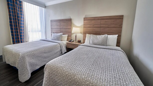 A hotel room with two beds, a nightstand with a lamp, and a window with curtains. - Two Double Beds - Standard (Fairhaven)