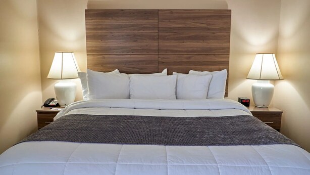 Habitación de hotel con una cama grande, dos mesitas de noche con lámparas, un cabecero de madera y un reloj digital.