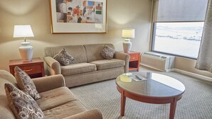 Habitación de hotel con sofá, sillón, una mesa de centro de vidrio, dos mesitas de noche con lámparas, un cuadro abstracto enmarcado y un ventanal con vistas.