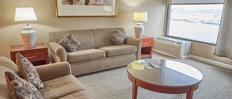 A hotel room with a sofa, armchair, a glass coffee table, two bedside tables with lamps, a framed abstract painting, and a large window with a view.