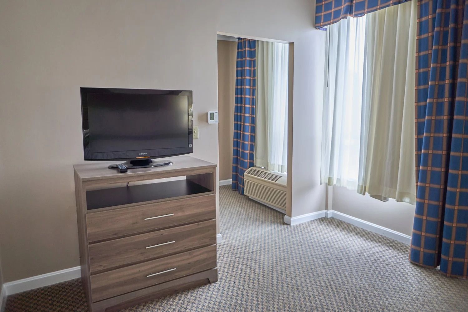 A hotel room with a flat-screen TV on a wooden cabinet, a window with curtains, and a radiator.