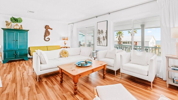 A spacious living room with a wooden floor, white sofas, a teal cabinet, and a balcony with a view of palm trees and houses.