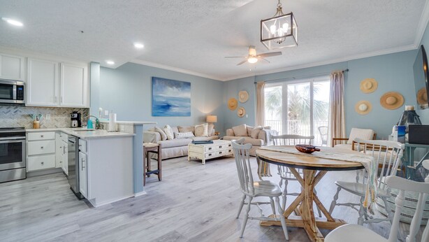 A modern kitchen with white cabinets, a microwave, and a dining area with a wooden table and white chairs. - Gorgeous condo with gulf views, pool & hot tub in Mexico Beach. Sleeps 6. (Mexico Beach)