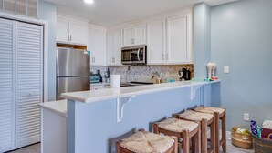 A kitchen with white cabinets, a stainless steel refrigerator, a microwave, and a bar area with wooden stools. - Gorgeous condo with gulf views, pool & hot tub in Mexico Beach. Sleeps 6. (Mexico Beach)