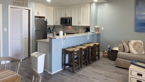 A modern kitchen with white cabinets, a stainless steel refrigerator, and a microwave. There is a small island with stools and a sink. The room has a gray wall and a beige sofa. - Gorgeous condo with gulf views, pool & hot tub in Mexico Beach. Sleeps 6. (Mexico Beach)