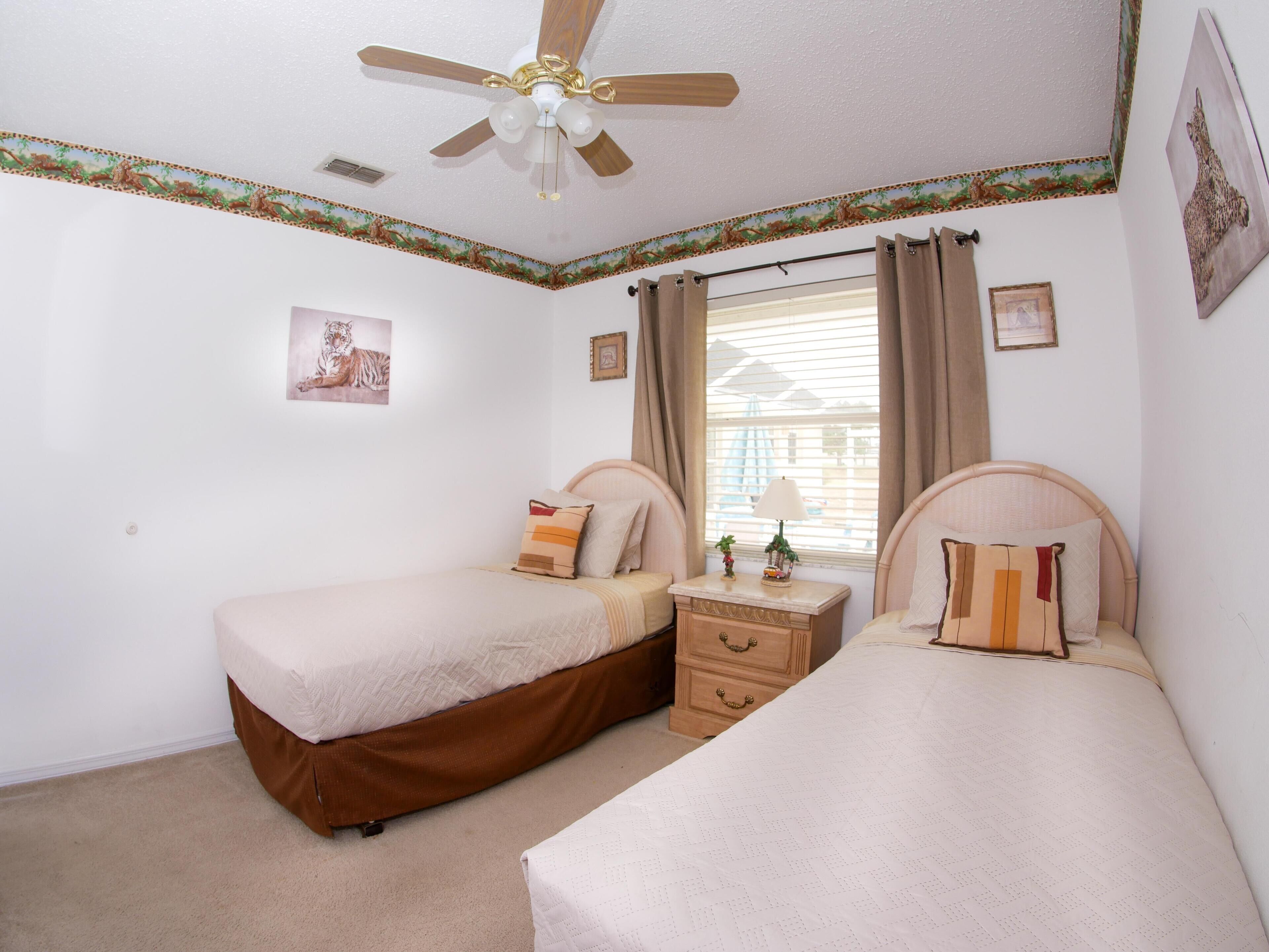 A bedroom with two beds, a ceiling fan, a window with curtains, and a small bedside table with a lamp.