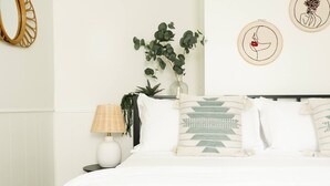 Une chambre à coucher bien rangée, avec un lit, des oreillers, une lampe de chevet, un vase avec des plantes et des œuvres d’art encadrées accrochées au mur.