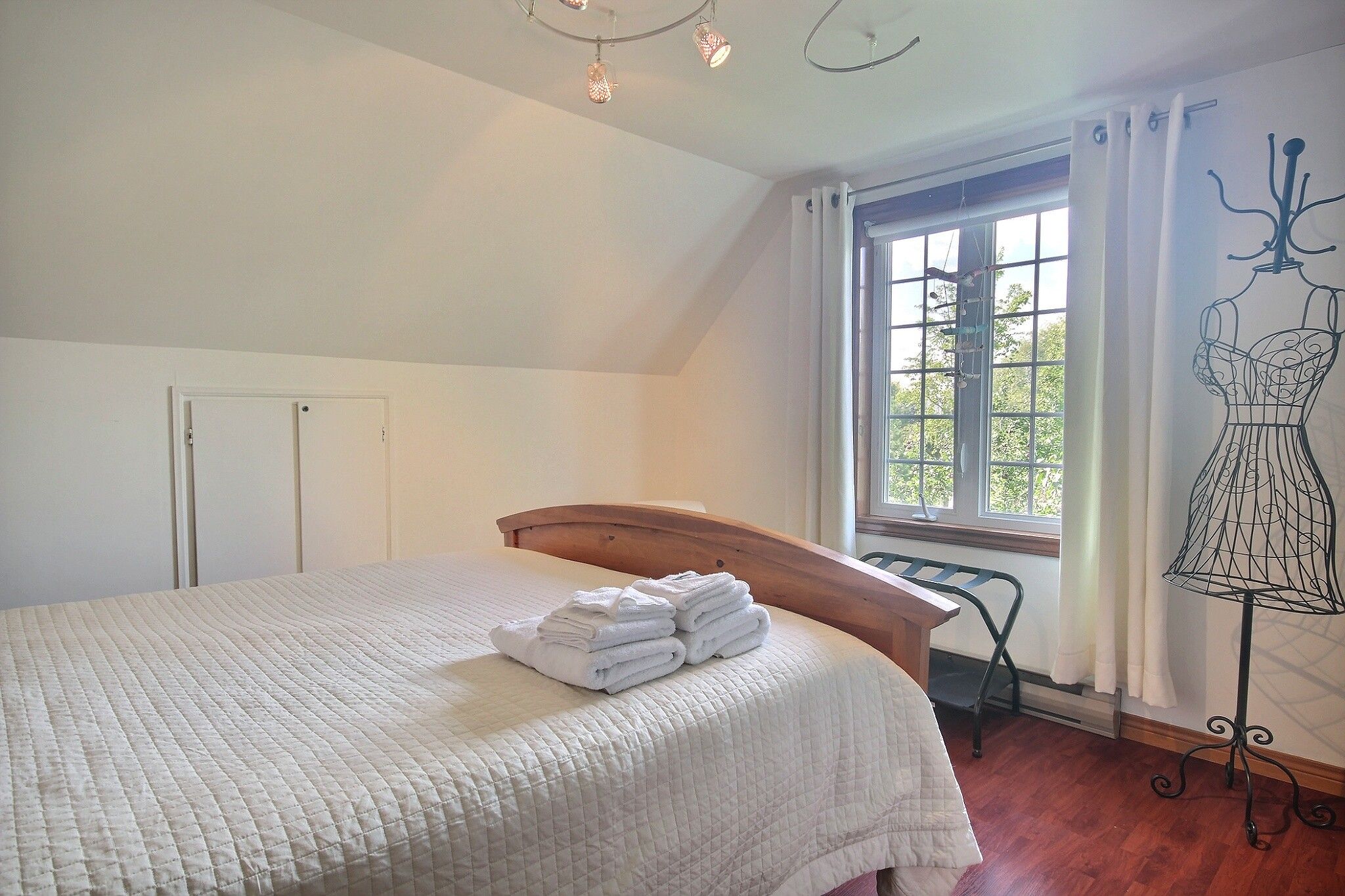Un dormitorio con cama, ventana con cortinas, una silla de metal y un maniquí.