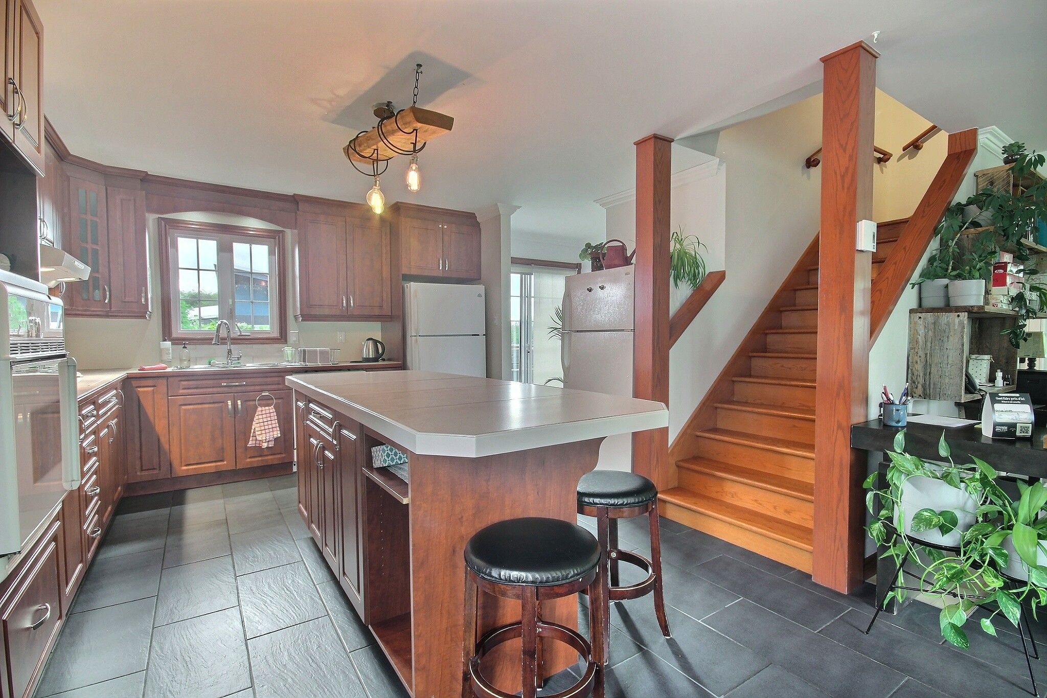 A kitchen with wooden cabinets, a central island with stools, a refrigerator, and a staircase leading to another level.