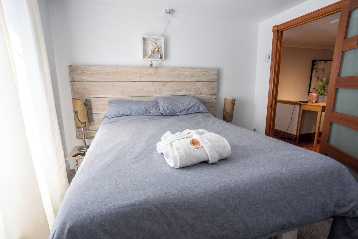 A neatly made bed with a grey bedspread and a white towel with a logo, a wooden headboard, a small wall-mounted light, and a wooden door with frosted glass panels.