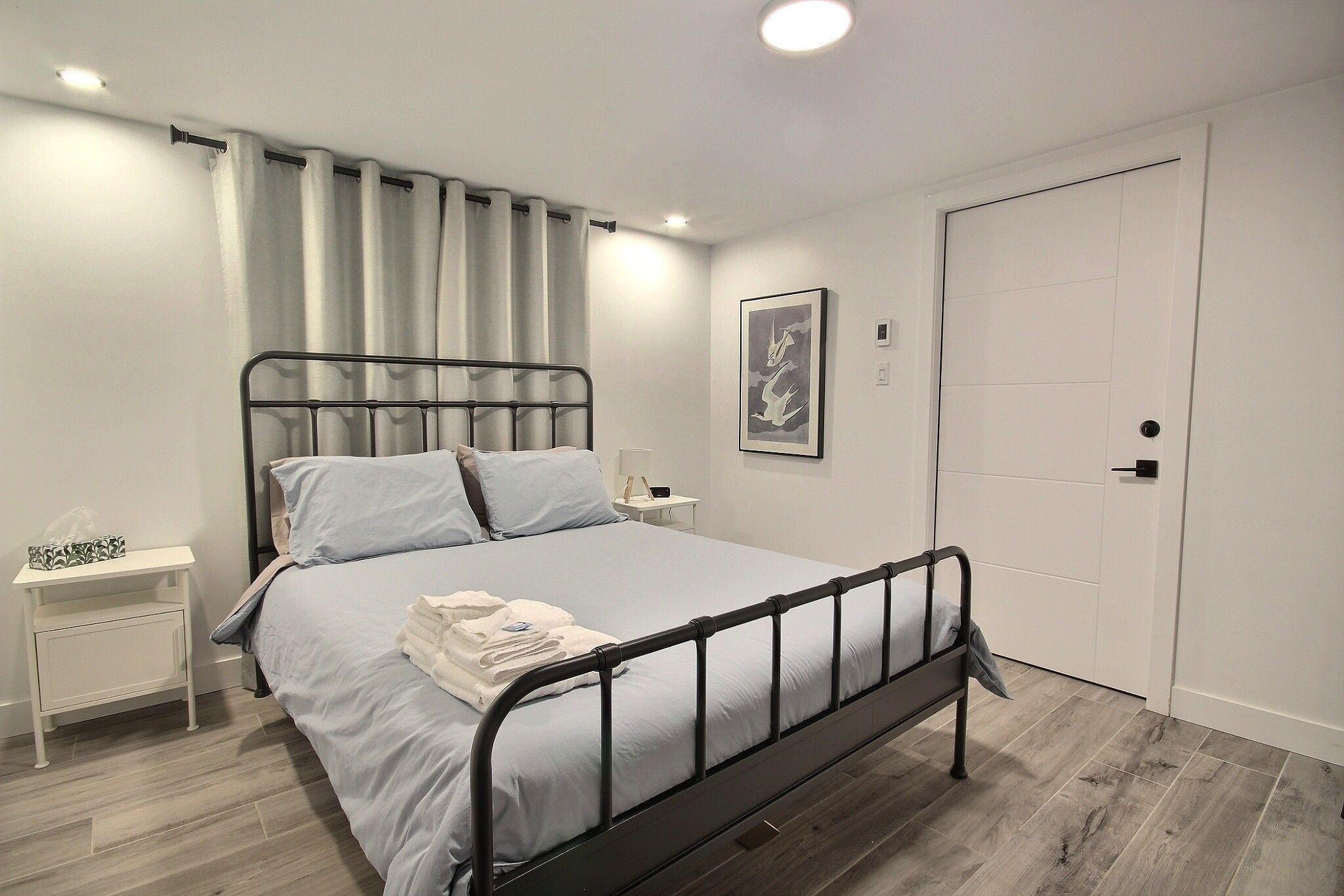Cama bem arrumada com estrutura de metal, mesa de cabeceira com luminária e porta à direita.