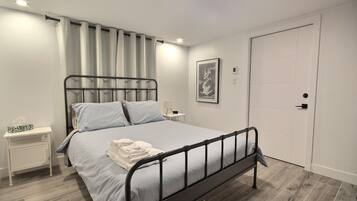 Cama bem arrumada com estrutura de metal, mesa de cabeceira com luminária e porta à direita.