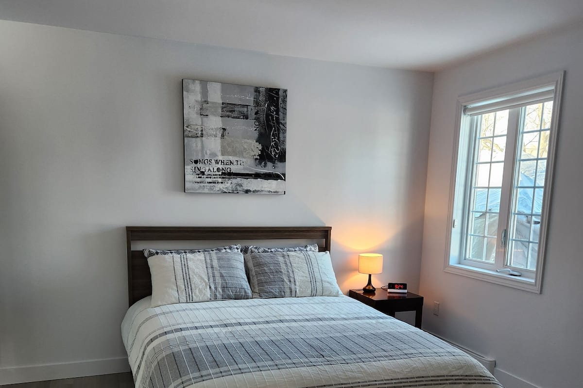 A bedroom with a bed, a bedside table with a lamp, a window, and a wall-mounted artwork.