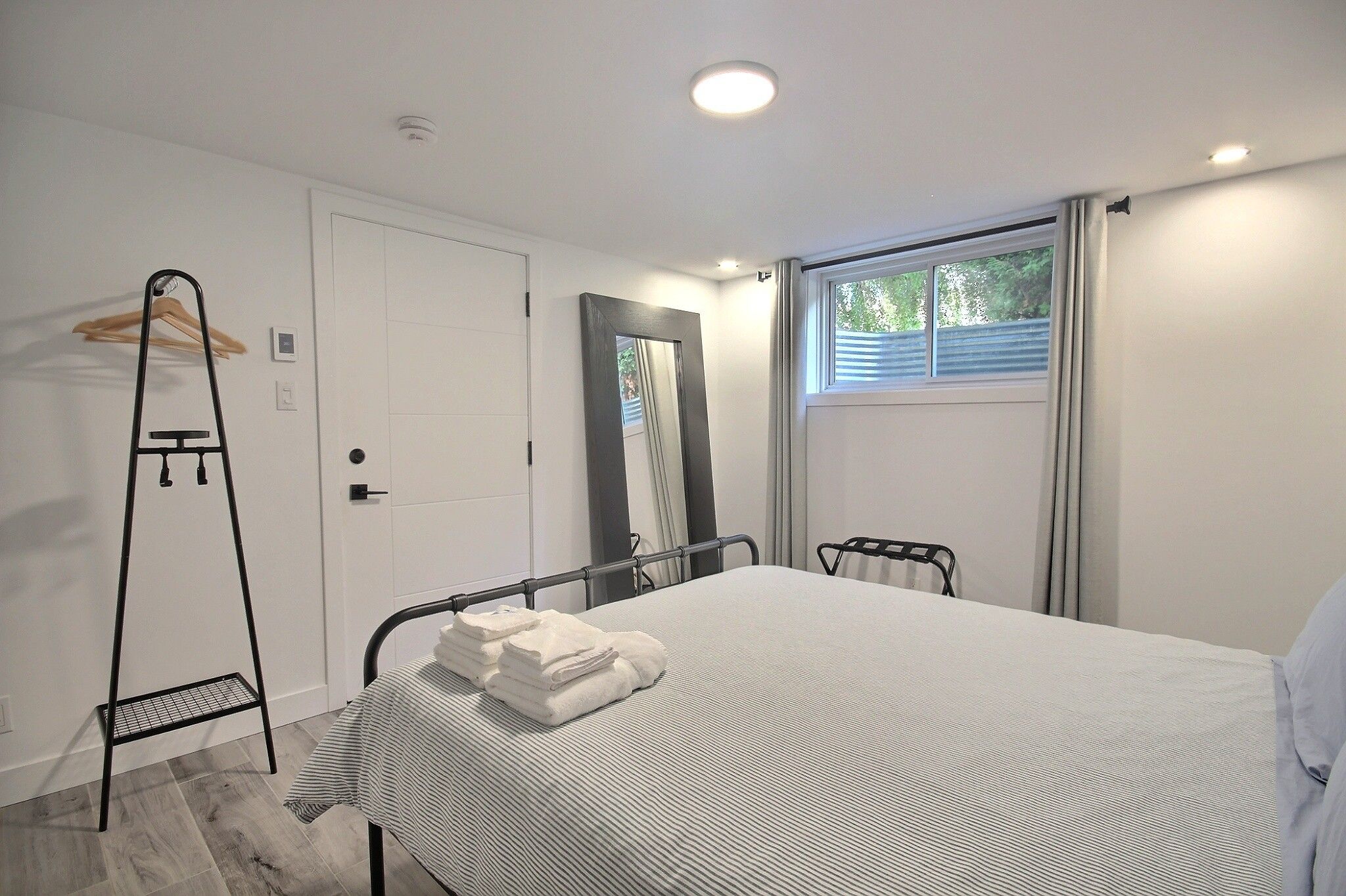 A bedroom with a bed, a bedside table, a hanging rack, a window with curtains, and a door.