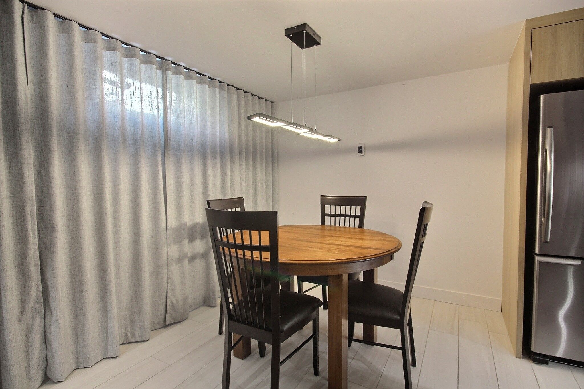 Een eetgedeelte met een ronde houten tafel, vier stoelen, een moderne hanglamp en een roestvrijstalen koelkast.