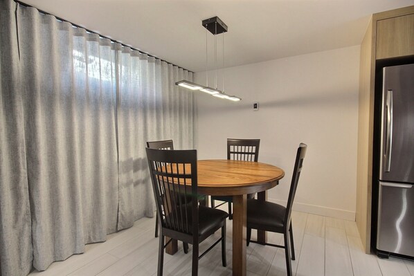A dining area with a round wooden table, four chairs, a modern pendant light, and a stainless steel refrigerator. - Aubergélit (Saint Jean Port Joli)