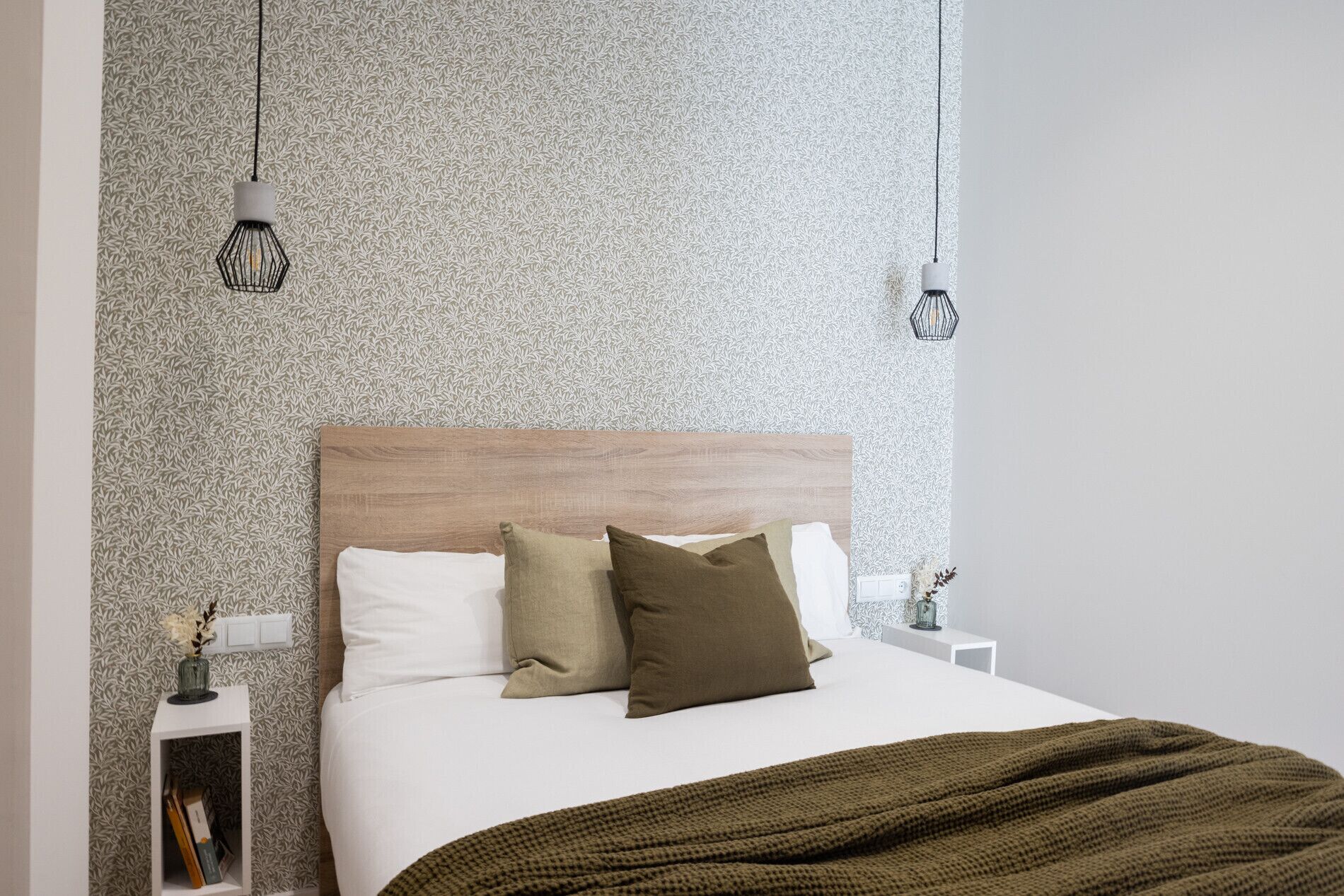 Une chambre avec un papier peint à motifs, une tête de lit en bois, un couvre-lit blanc et un plaid vert. On y trouve deux tables de chevet ornées d’objets décoratifs et deux lampes suspendues.