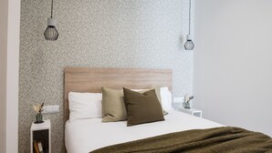 A bedroom with a patterned wallpaper, a wooden headboard, a white bedspread, and a green throw blanket. There are two bedside tables with decorative items and two hanging light fixtures. - The Sea Rooms VIII | AC & WIFI | 15 min Beach (Valencia)