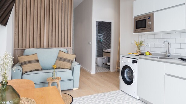 A modern apartment with a living area featuring a light blue sofa, a wooden slatted wall, and a small wooden table with a vase. The kitchen includes a washing machine, a microwave, and a sink with a tiled backsplash. - The Sea Rooms VIII | AC & WIFI | 15 min Beach (Valencia)
