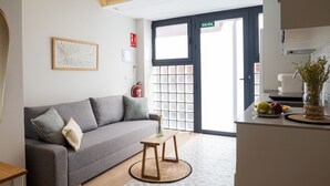 Un salon moderne avec un canapé gris, une petite table en bois et un coin cuisine agrémenté d’un panier de fruits et d’un vase.