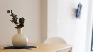 Una mesa de madera con un florero, un banco y una silla en una habitación con suelo de baldosas y pared blanca.