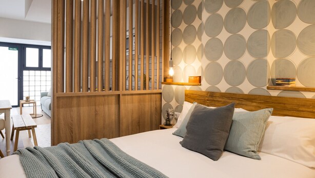 Un dormitorio con cabecera de madera, una cama con varios almohadones, una mesita de noche con lámpara y una pared con diseños circulares.