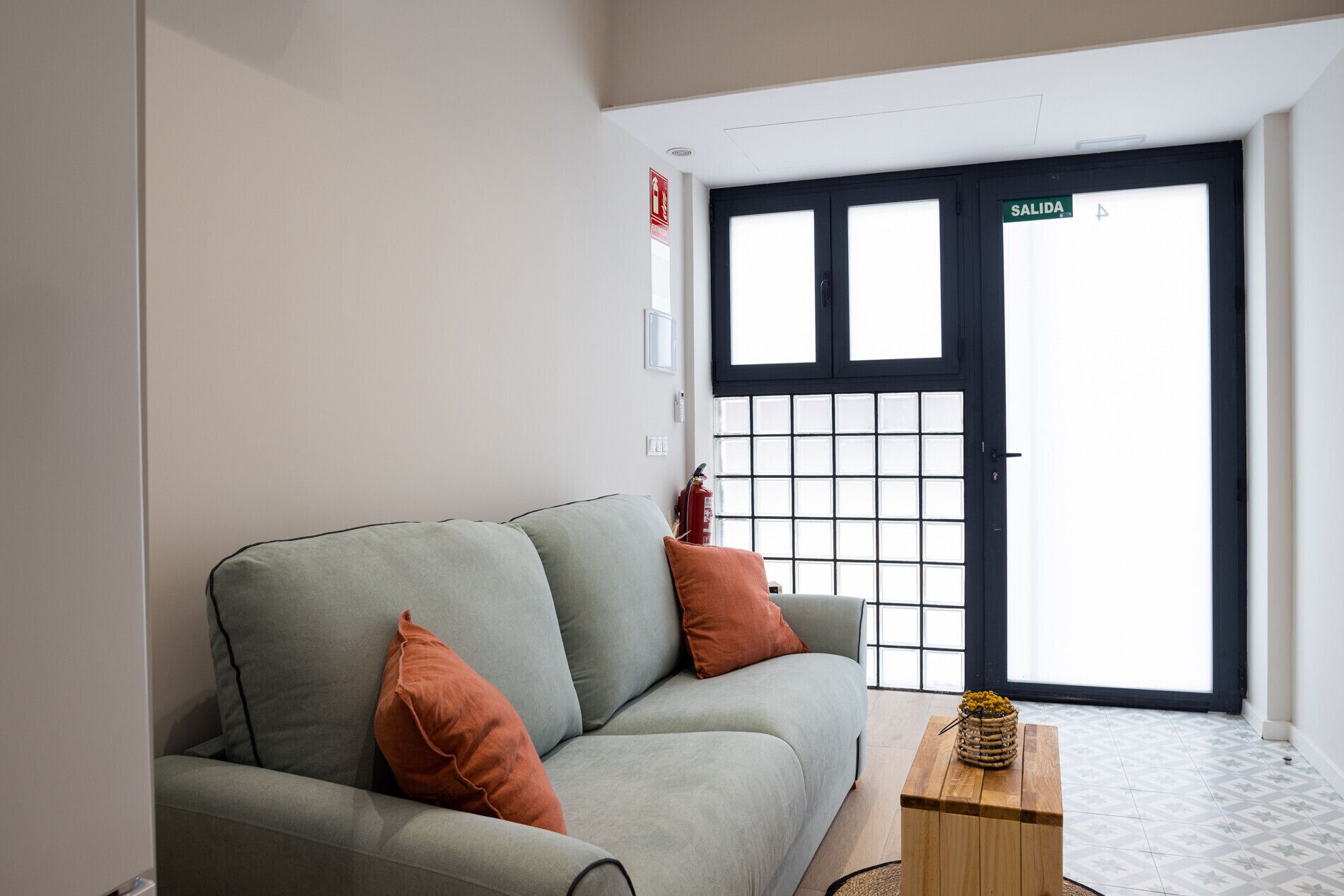 A modern living room with a grey sofa, two orange cushions, a wooden side table with a decorative item, and a black door with a grid pattern.