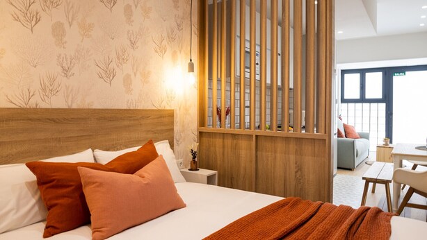 A hotel room with a bed, a wooden headboard, a nightstand, a desk, and a view of the outside.