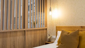 Habitació d'hotel amb capçal de llit de fusta, una finestra gran amb persianes verticals, una taula de nit amb una làmpada i un llit amb llençol marró.