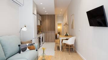 Un espacio habitable compacto con sofá, una mesa de comedor, un televisor montado en la pared y una zona de cocina visible a través de una puerta abierta.