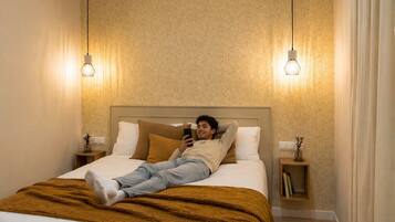 Una persona recostada en una cama dentro de una habitación con papel tapiz estampado, dos lámparas de noche y una mesita de noche con un libro.