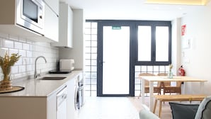 Una cocina moderna con armarios blancos, un microondas, un lavabo y una lavadora. Hay una zona de comedor con una mesa y sillas, y una puerta señalizada con 