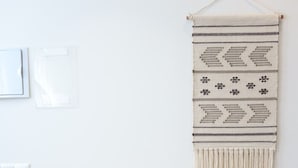 Una habitación con una mesa de madera, un tapiz de macramé en la pared y un jarrón pequeño con flores secas sobre la mesa.