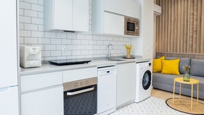 Een moderne keuken met witte kastjes, een ingebouwde magnetron, een vaatwasser en een wasmachine. In de aangrenzende woonkamer staat een grijze bank met gele kussens.