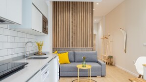 Een moderne keuken met witte kasten, een roestvrijstalen spoelbak en een ingebouwde oven. Een grijze bank met gele kussens staat voor een wand van houten latten. Een kleine ronde tafel met een geel onderstel is voor de bank geplaatst.