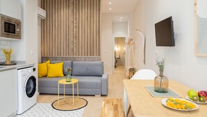 Een modern appartement met een grijze bank, gele kussens, een klein rond tafeltje, een wasmachine, een eettafel met fruit en een aan de muur bevestigde televisie.