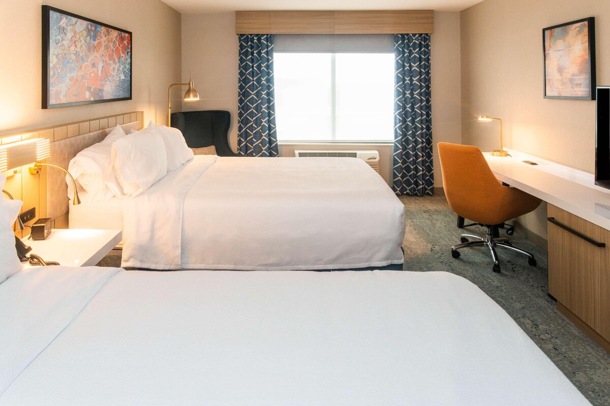 A hotel room with two beds, a desk, a chair, and a large window with curtains.