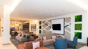 Eine moderne Hotelhalle mit Sitzgelegenheit, einem an der Wand befestigten Flachbildfernseher und einer Küche im Hintergrund.