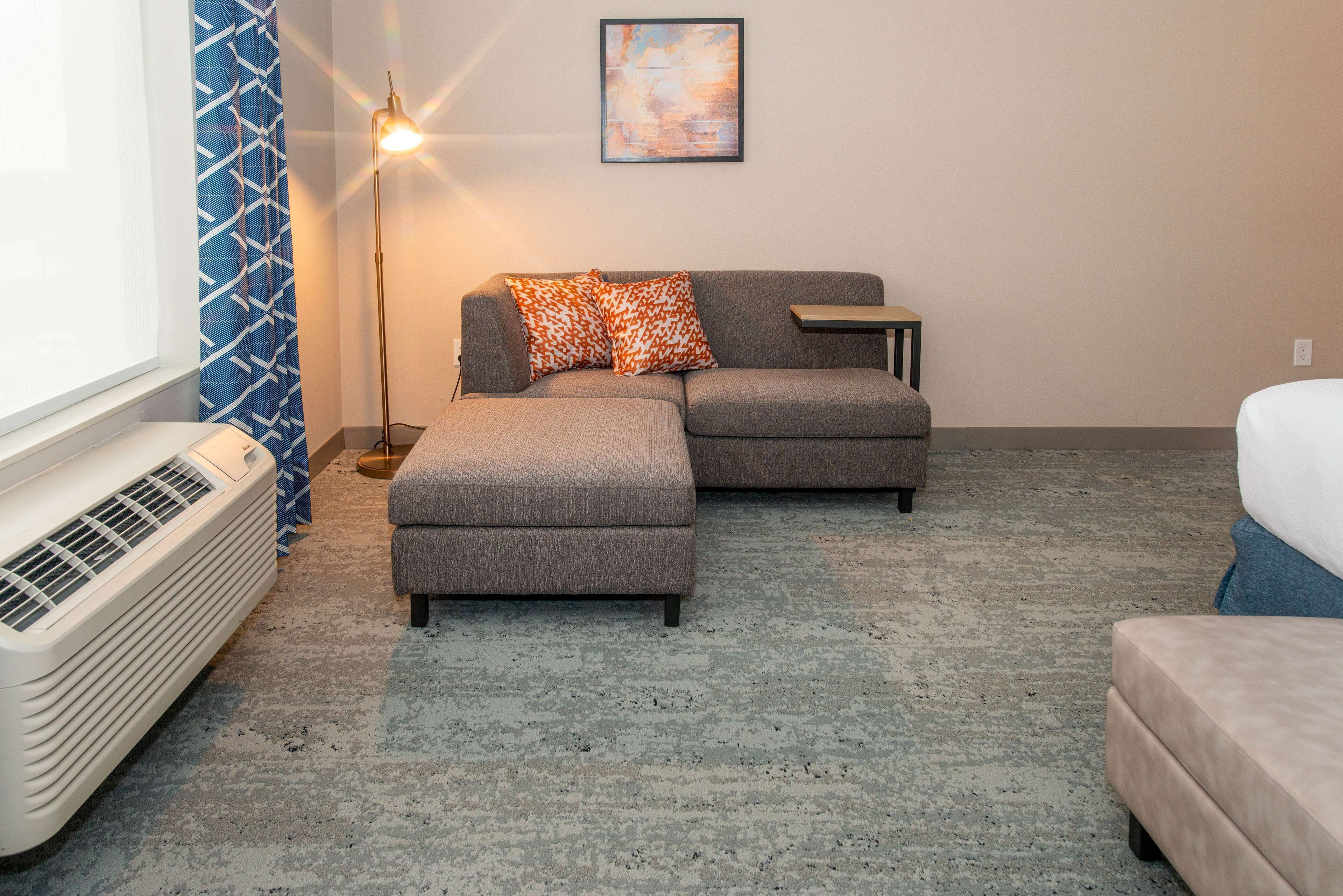 A modern living room with a sectional sofa, an ottoman, a standing floor lamp, and a large window with patterned curtains.