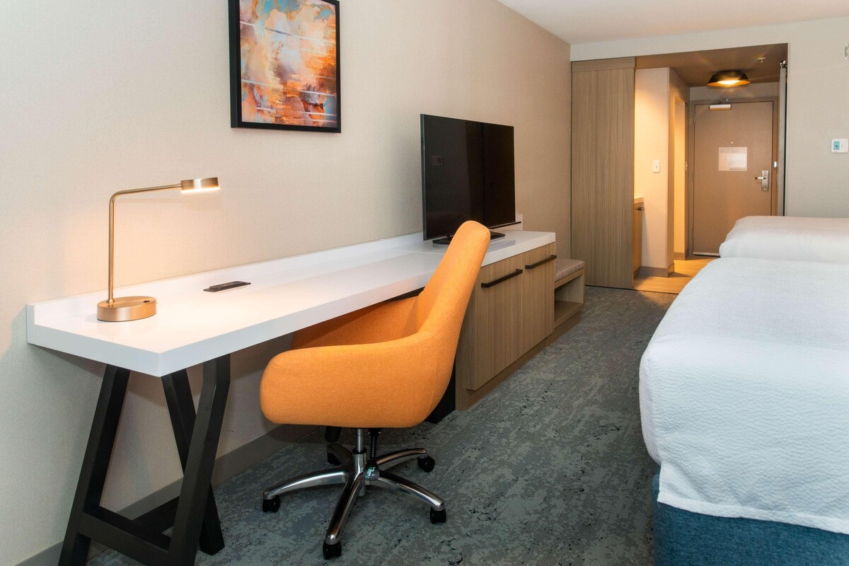 A hotel room with a white desk, an orange chair, a flat-screen TV, a bed, and a door leading to another room.