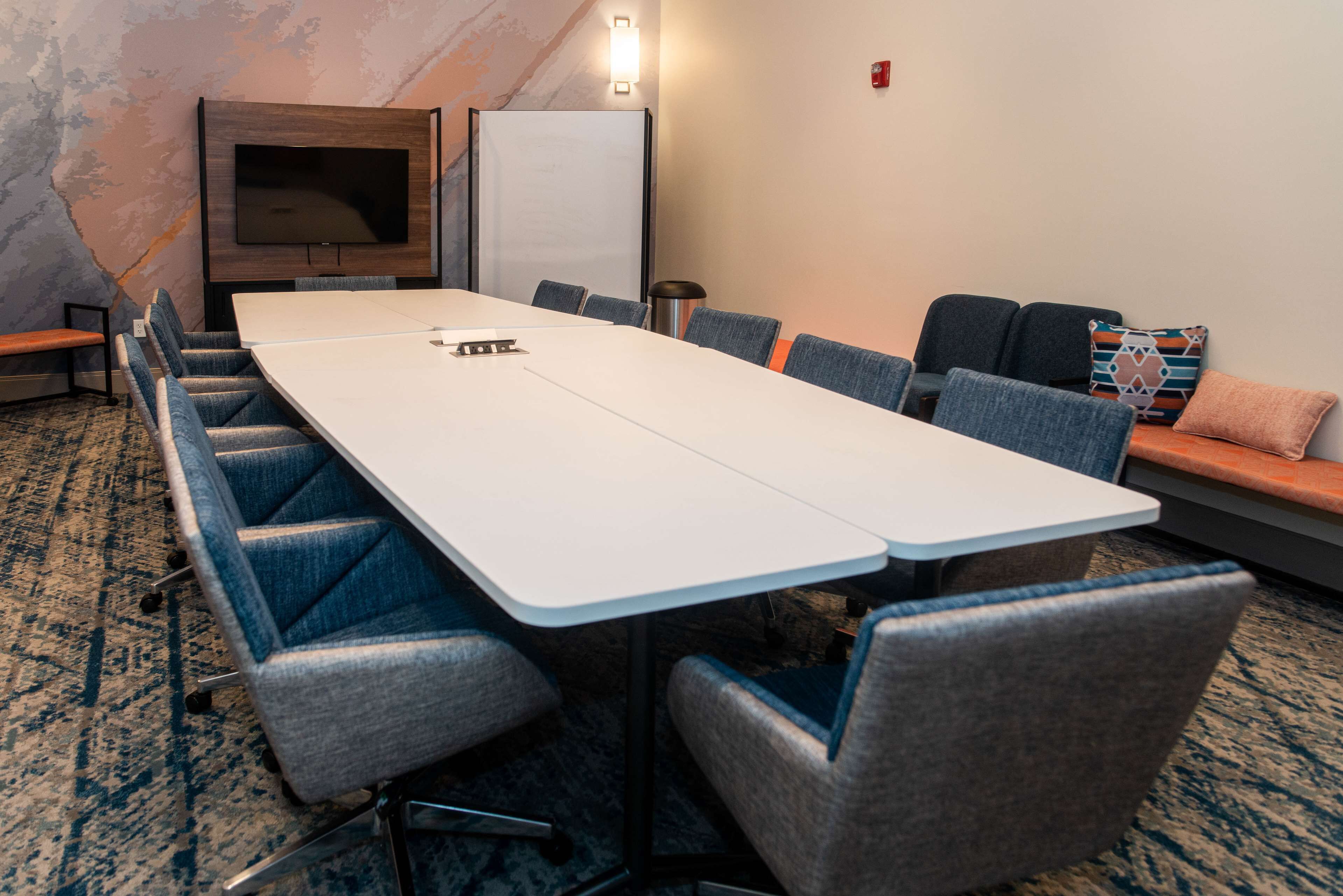 A conference room with a long table, blue upholstered chairs, a flat-screen TV, and a whiteboard.
