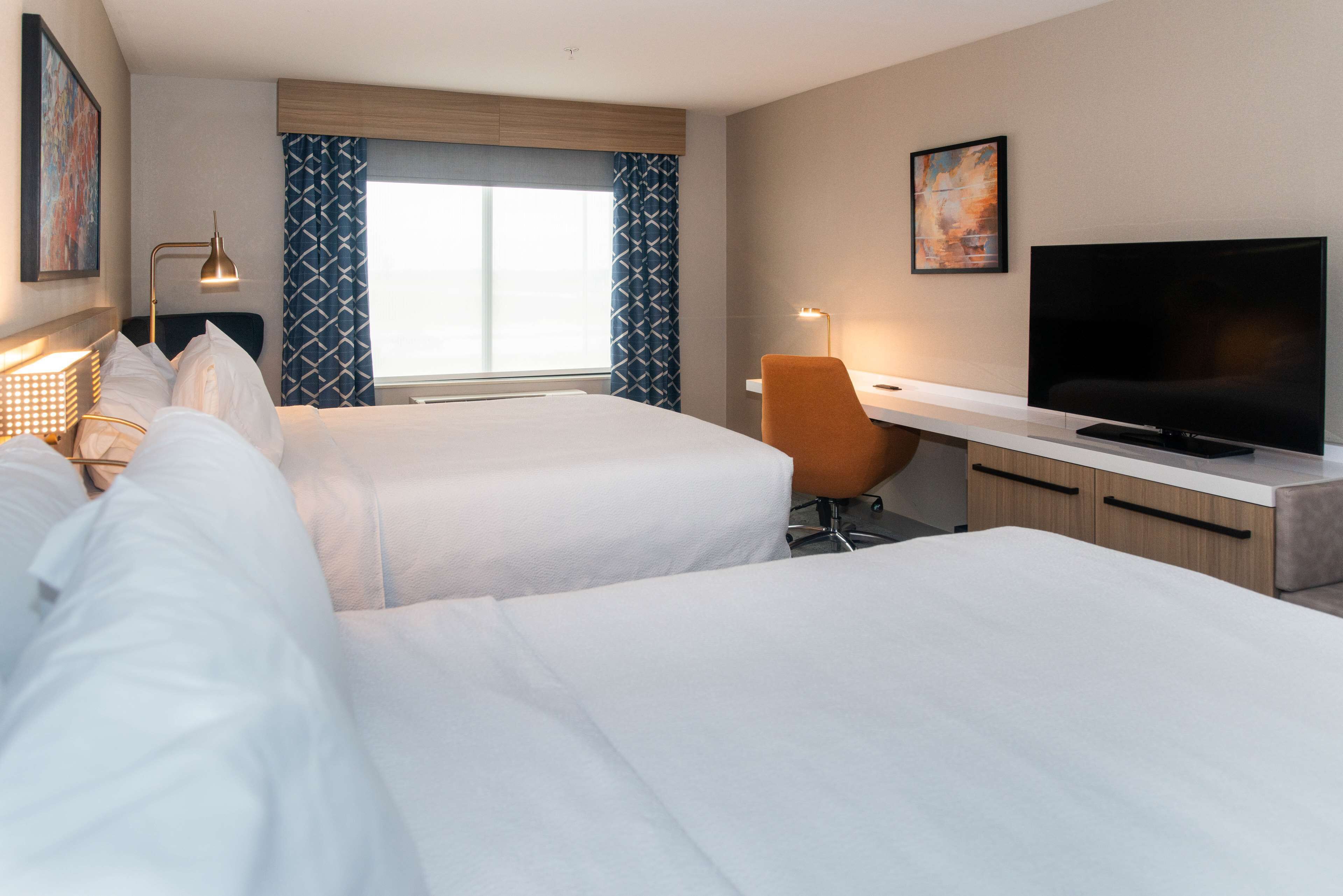 A hotel room with two beds, a desk, a TV, and a window with patterned curtains.