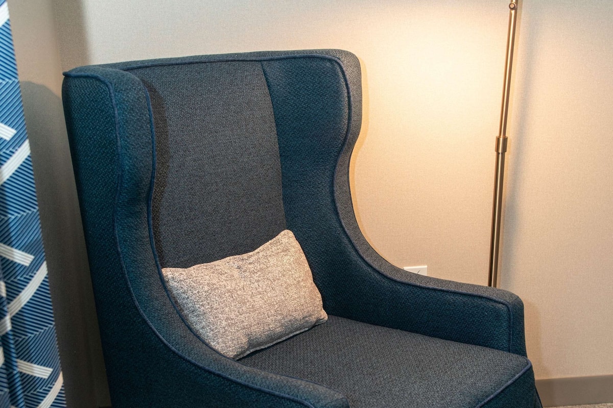 A blue armchair with a cushion, a round wooden table, and a floor lamp in a room with a plain wall and a patterned curtain.