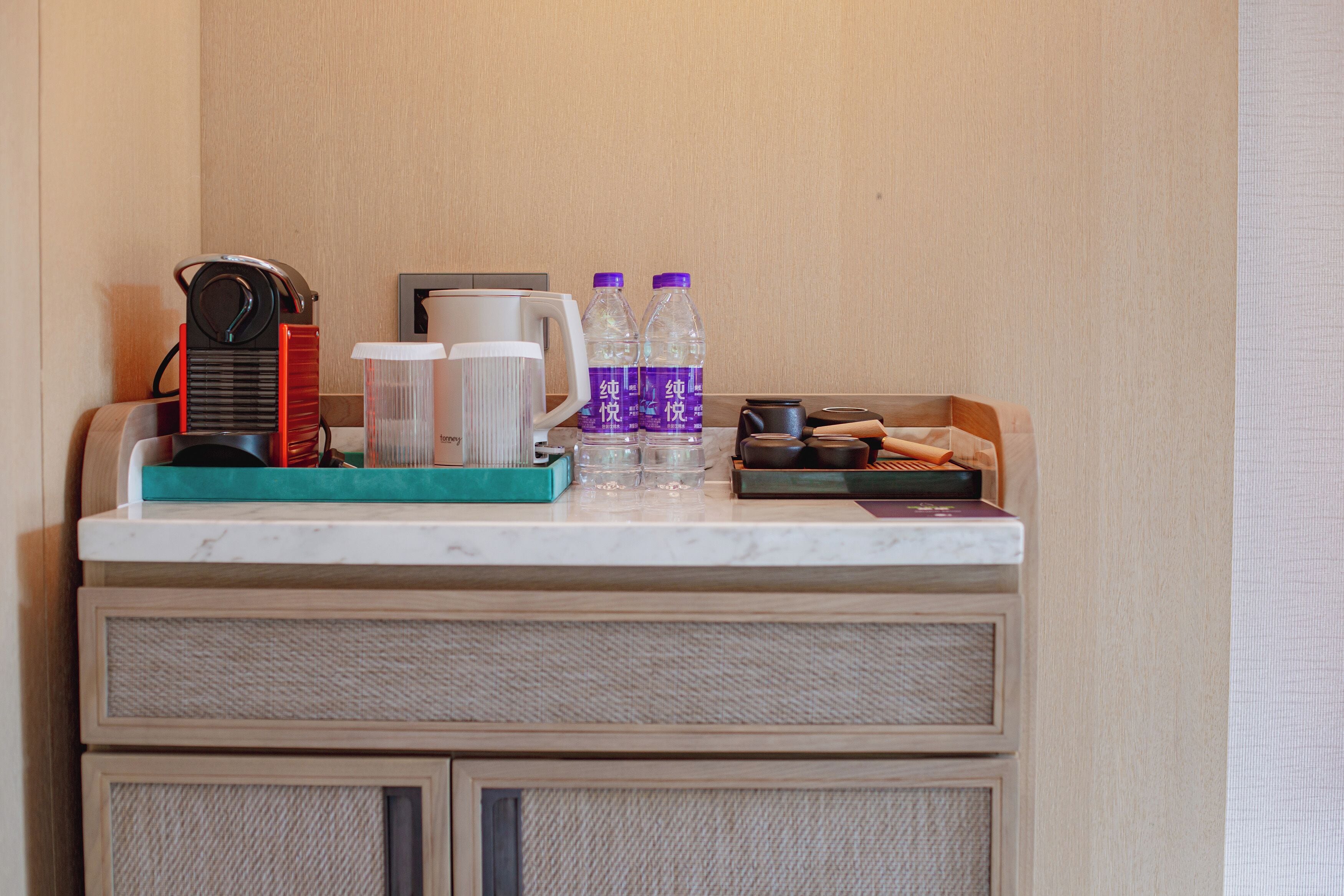 Un minibar de hotel con una cafetera, dos botellas de agua, una jarrón y una bandeja con vasos y una caja de pañuelos.